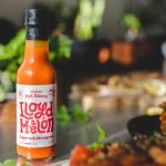 Flaske med Lloyd & Melon hot sauce med cayenne og sitrongress, satt på et bord med friske urter og mat i bakgrunnen.