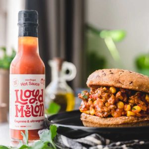 Flaske med Lloyd & Melon Hot Sauce ved siden av en saftig burger fylt med krydret plantebasert fyll og mais, servert på en svart tallerken.