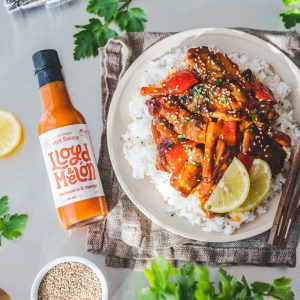 En tallerken med ris toppet med karamellisert kylling og grønnsaker i en spicy saus, servert med limebåter og sesamfrø. Ved siden av ligger en flaske Lloyd Melon Habanero & Melon hot sauce.