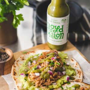 En saftig taco med pulled chicken, guacamole, rødløk og koriander, toppet med Lloyd Melon Jalapeño & Ananas hot sauce. Flasken står i bakgrunnen på en rustikk trefjøl.