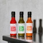 Tre flasker Lloyd Melon hot sauce i forskjellige farger – rød, grønn og oransje – står oppstilt på en stålbenk mot en betongvegg.