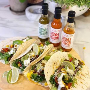 Tre sauser fra Lloyd Melon i ulike smaker står bak en fargerik servering av tacos med ferske grønnsaker, lime og revet ost, servert på en trefjøl."