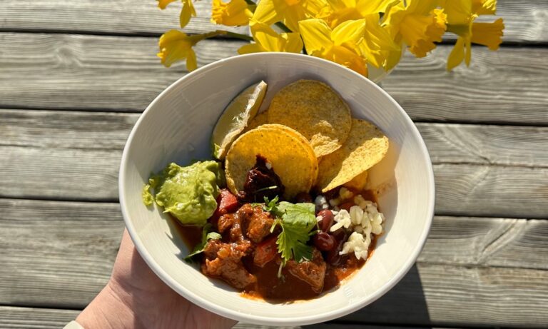 Skål med sjokolade-lam-chili toppet med koriander og tortillachips, servert i en grå bolle på høstfarget bakgrunn.
