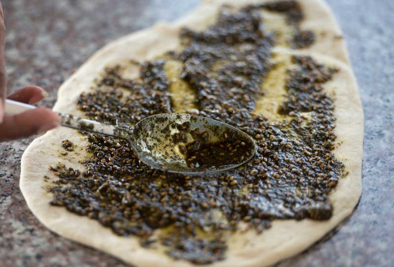 En hånd smører za’atar-olje over et ferskt manakish-brød før steking. Tradisjonell levantinsk bakst med en aromatisk krydderblanding.